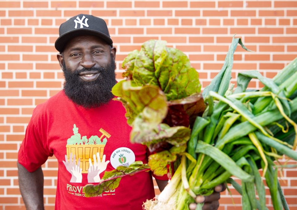 Hamadi Ali dressed in red PFC shirt and holding an assortment of fresh vegetables